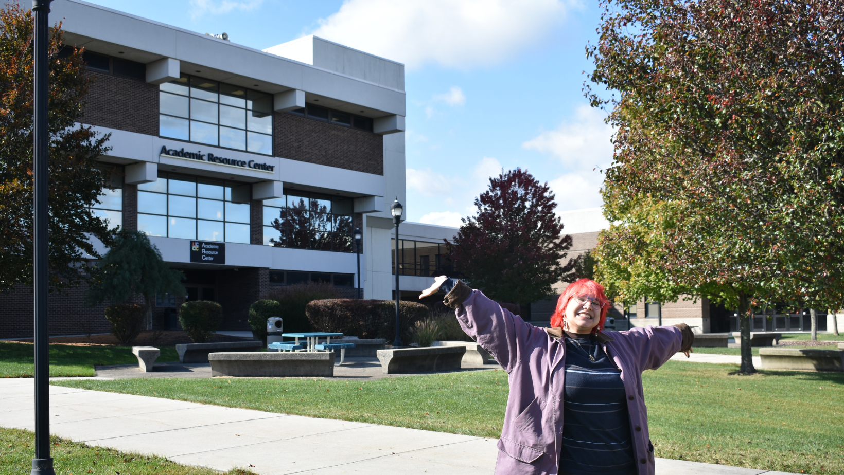Image shows Alex Maaser smiling with their arms raised in front of the ARC on main campus.