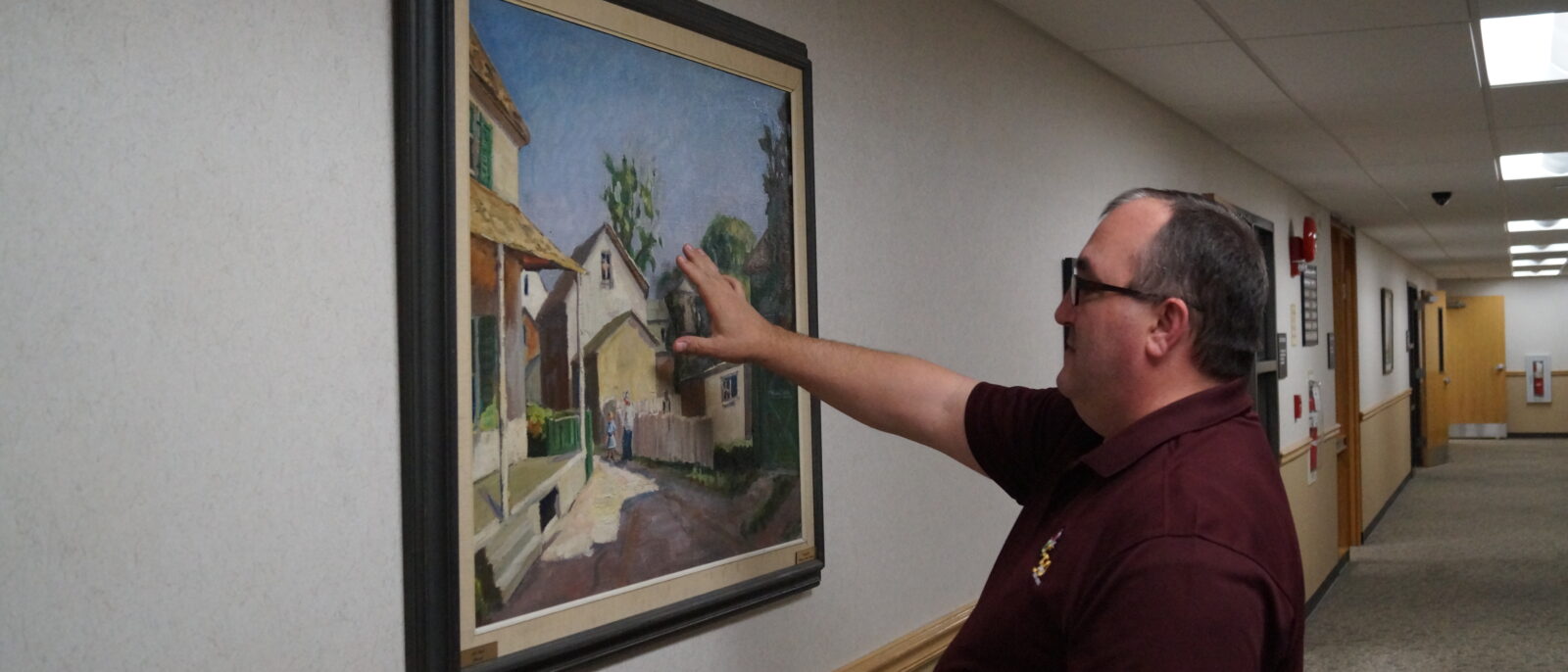 A man in a maroon LCCC polo shirt holds his hand up to the front of a painting in a hallway.