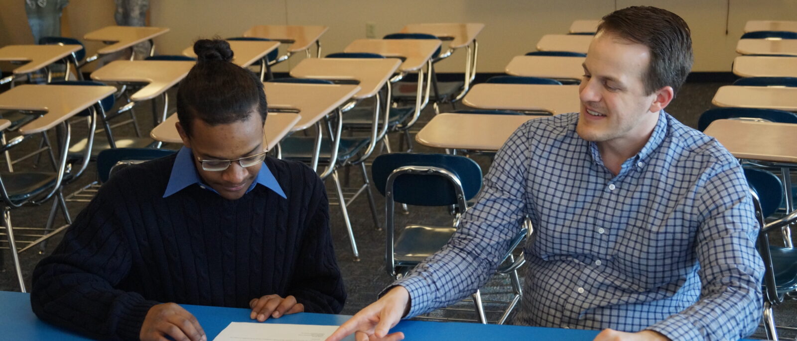 A teenage student wearing glasses looks at a paper while a teacher in a checkered dress shirt sits next to him and points at the paper.