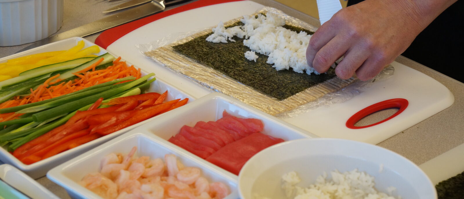 Shallow dishes filled with ingredients such as peppers, shrimp and rice are on a table while two hands work on putting rice on seaweed paper.
