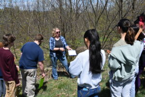 Image shows a representative from Trexler Game Preserve holding a clipboard and explaining processes to students while at a tree grove.
