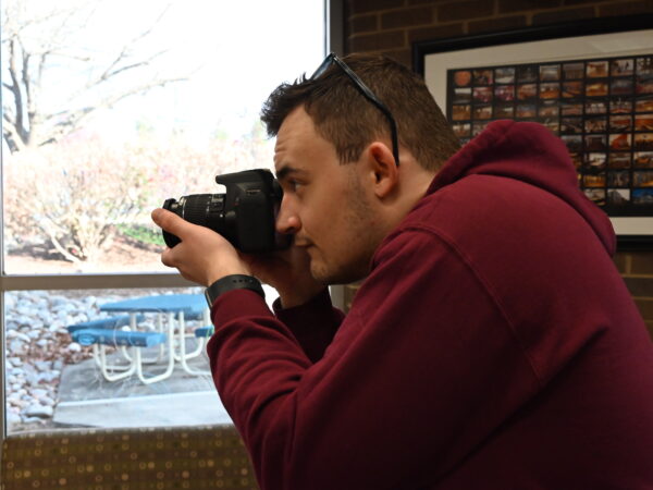 Image shows Brody Owens holding a camera up to his face, taking a photo in the Rothrock Library in front of a window.