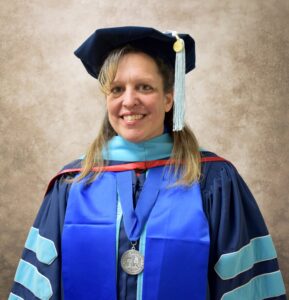 A woman in full doctoral regalia smiles in front of a photography backdrop.