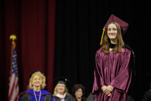 Image shows Diana Moussa in graduation regalia standing on the Commencement stage with the platform party behind her.