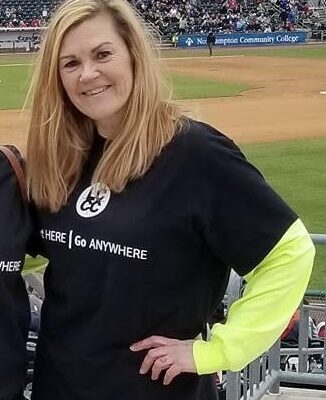 A woman with long blonde hair poses at a baseball game wearing a black Lehigh Carbon Community College t-shirt.