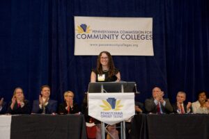 A panel of people sit and clap behind a young woman standing at a podium. At the podium and on a banner hanging behind everyone is the logo and text that reads "Pennsylvania Commission for Community Colleges"