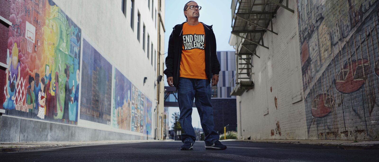 Jose Rivera stands in the middle of an alley wearing an orange t-shirt that reads "End Gun Violence Now".