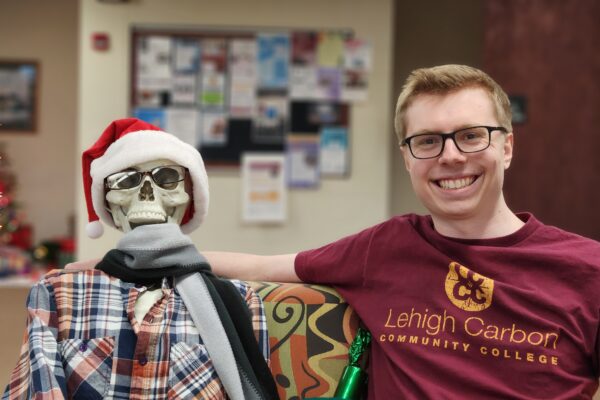 A young man wearing glasses and a Lehigh Carbon Community College t-shirt sits next to a skeleton dressed in winter attire and a santa hat in the Rothrock Library.